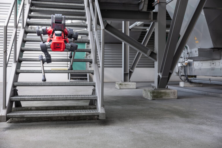Autonomous four-legged inspection robot climbing industrial metal stairs in a manufacturing facility.